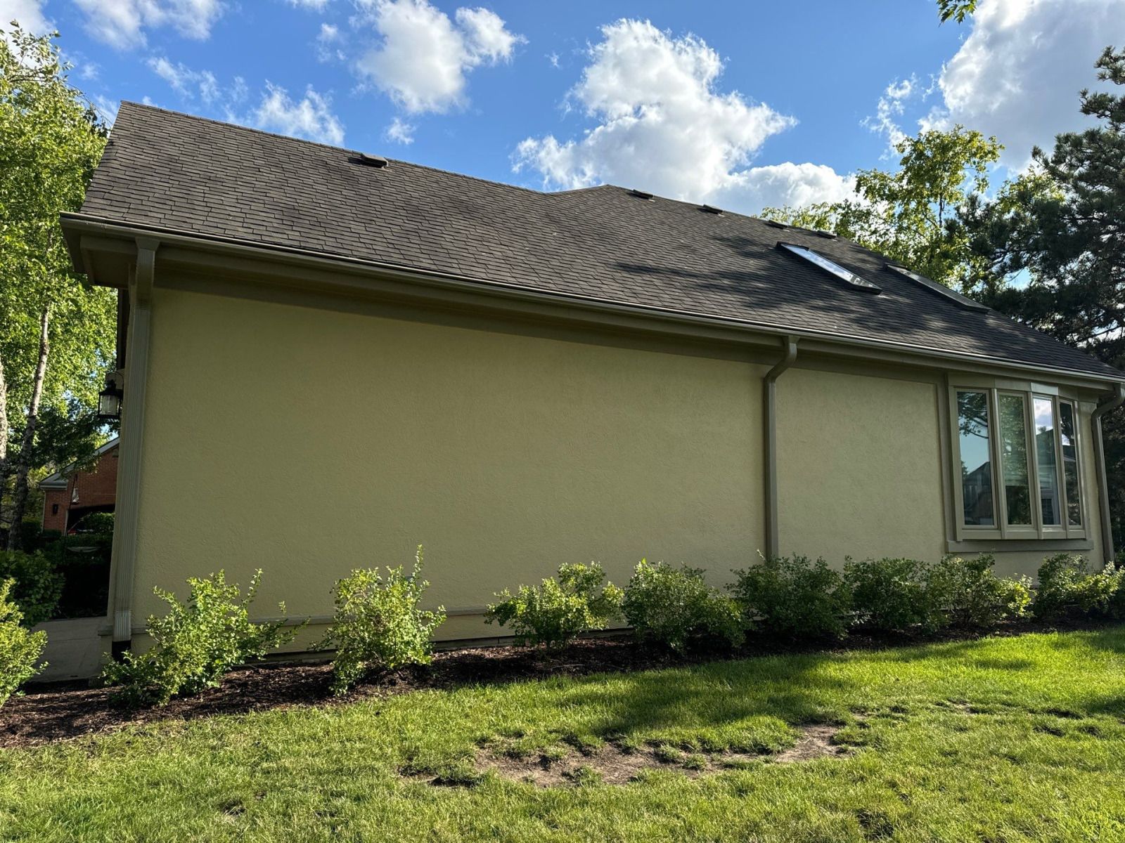 Single-story beige house with a dark shingled roof, small shrubs, and a green lawn under a blue sky.
