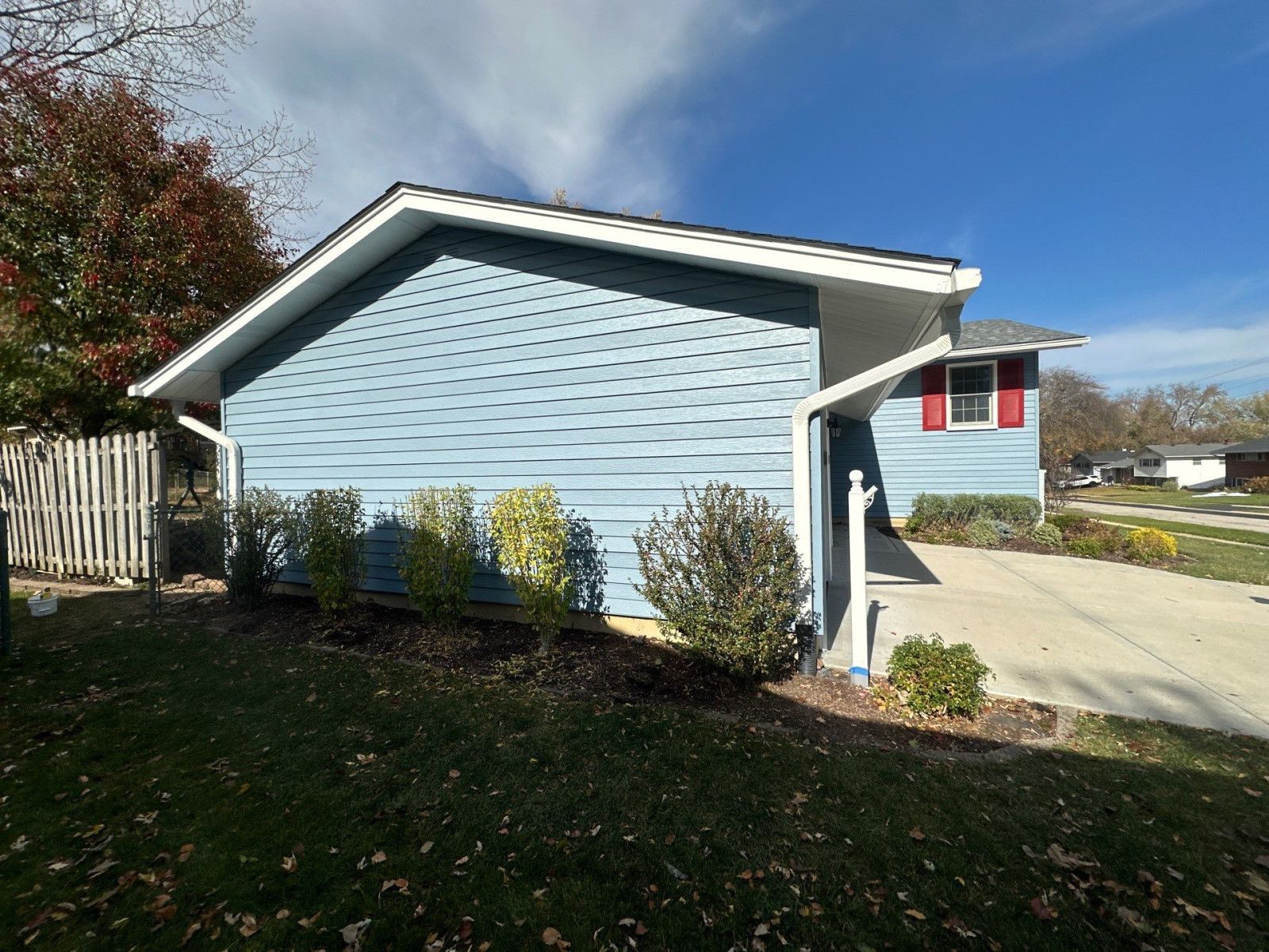 Blue house exterior with a driveway, small shrubs, and a tree in a sunny yard