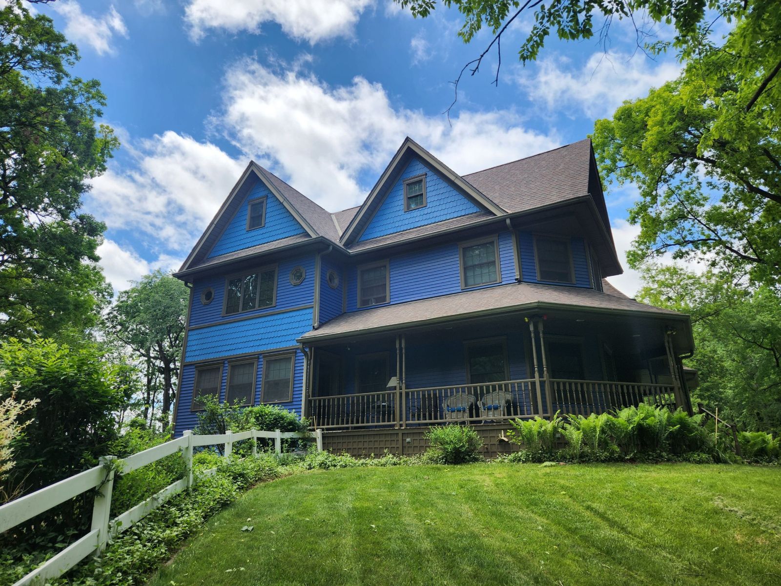 Blue Victorian house on a grassy hill with a porch, white fence, and trees under a cloudy sky