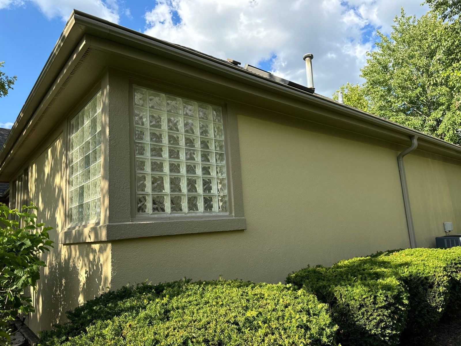 Side view of a beige house with glass-block window and green hedges under a sunny sky