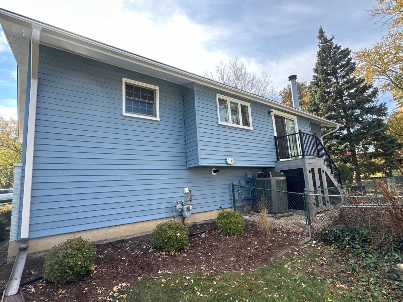 Blue two-story house exterior with basement windows, small porch stairs, and surrounding shrubs in a wooded yard