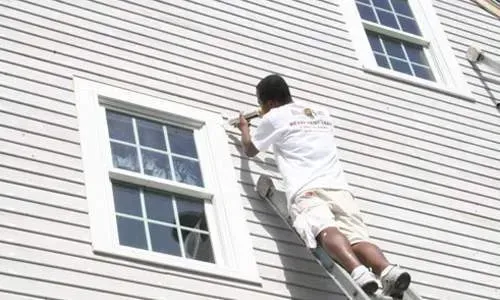Worker cleaning a white house exterior on a ladder near two windows
