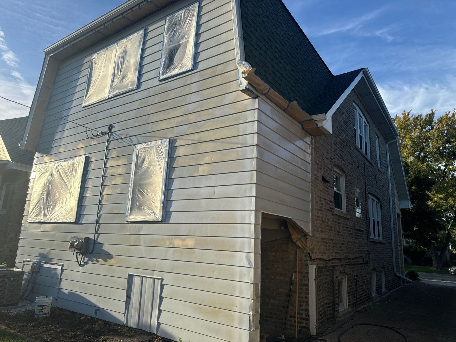 Side of a gray house with boarded windows and brick lower walls, viewed from an angle outdoors