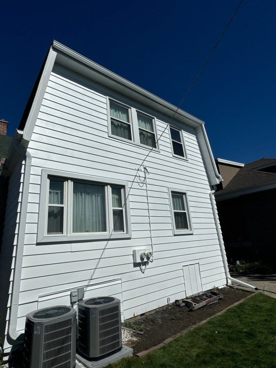 White two-story house with vinyl siding, two windows, and air conditioning units beside a narrow yard.