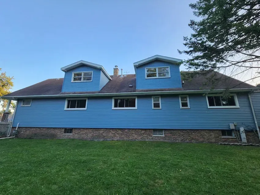 Blue house with two dormer windows, stone foundation, and a grassy backyard under a clear sky