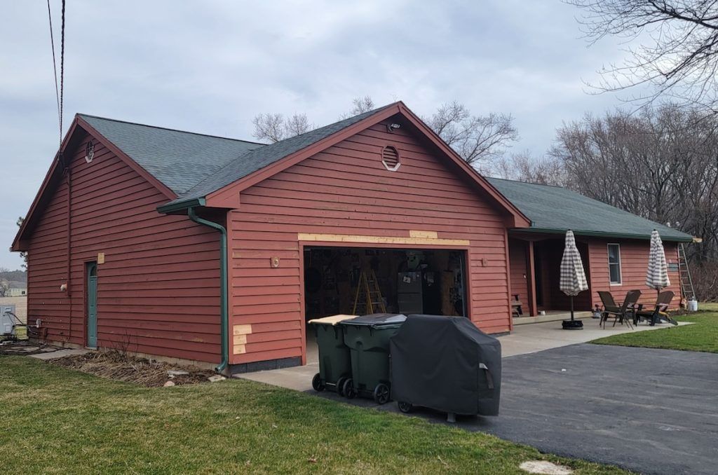 Red barn-style house with open garage, covered grill, and trash bins on a driveway