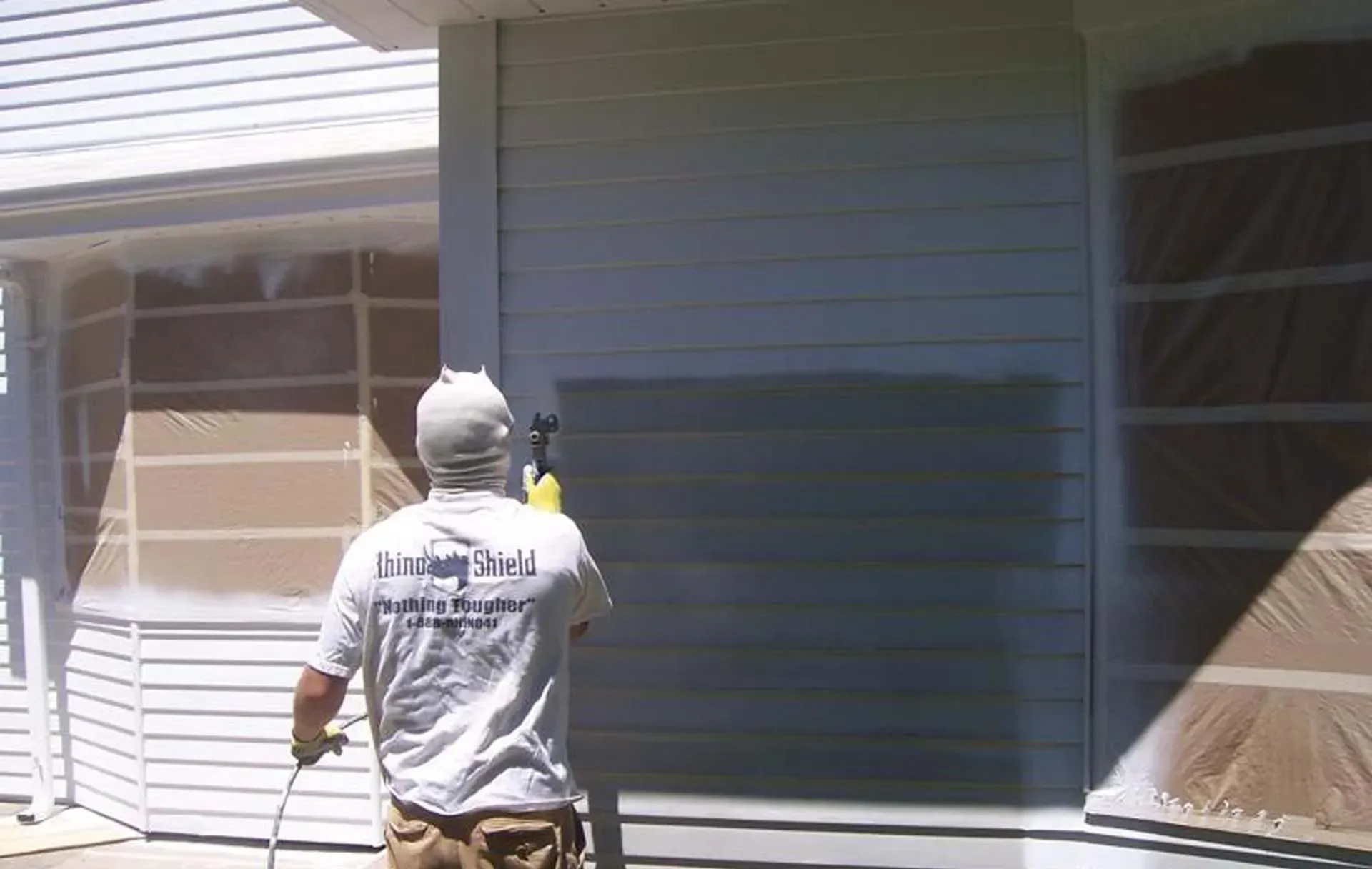 Person painting a house exterior gray next to a taped window in bright sunlight