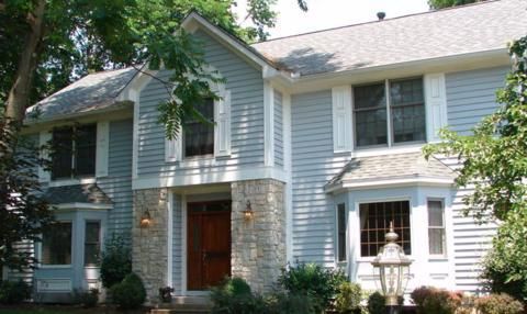 Two-story white house with dark shutters, a red front door, and a small front porch surrounded by trees and shrubs