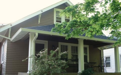 Small brown house with a front porch, white trim, and leafy trees in front