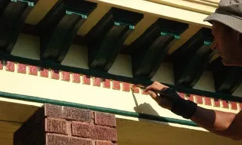 Person painting trim beneath roof eaves with a brush near red brick wall