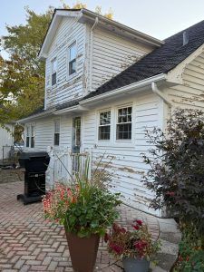 White two-story house with black roof, potted flowers, and a grill on a brick walkway