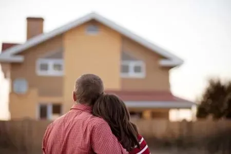 Couple standing together, seen from behind, looking at a tan house in a suburban yard.