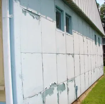White weathered building wall with peeling paint and small teal windows beside a grassy strip