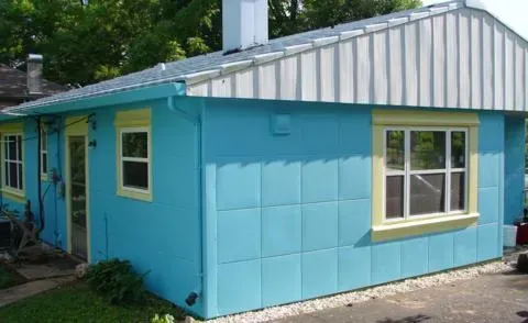 Light blue house exterior with white trim and a front window, seen beside a wooded yard