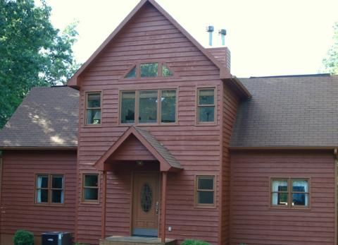 Red wooden house with a steep roof, front porch, and many windows surrounded by trees.