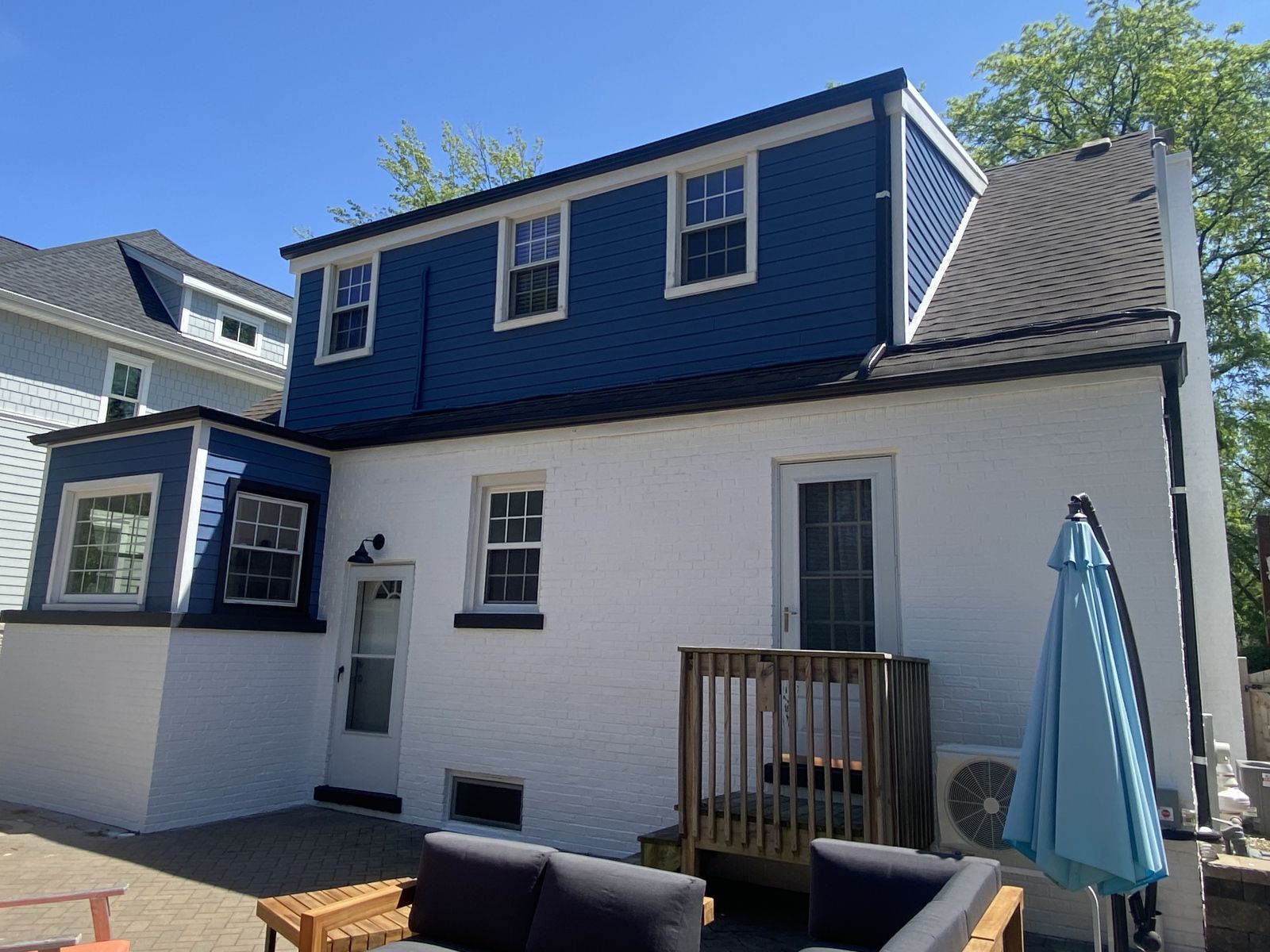 Blue-and-white two-story house with a small deck, closed umbrella, and adjacent outdoor patio area
