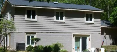 White two-story house with gray siding, dark roof, and windows amid greenery on a sunny day