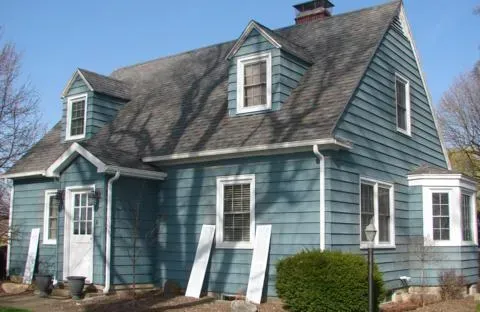 Blue-gray house with white trim, dormer windows, and shutters under a steep shingled roof