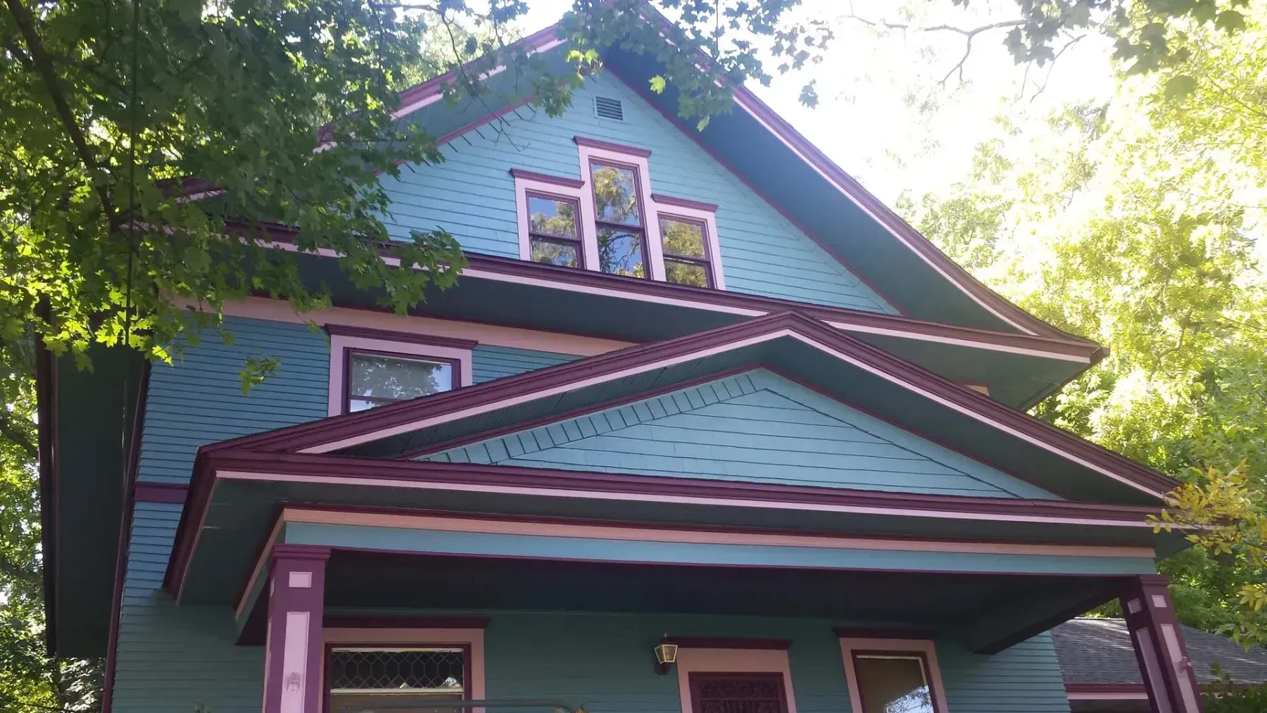 Victorian house with teal siding, purple trim, and a red-roofed porch under leafy trees