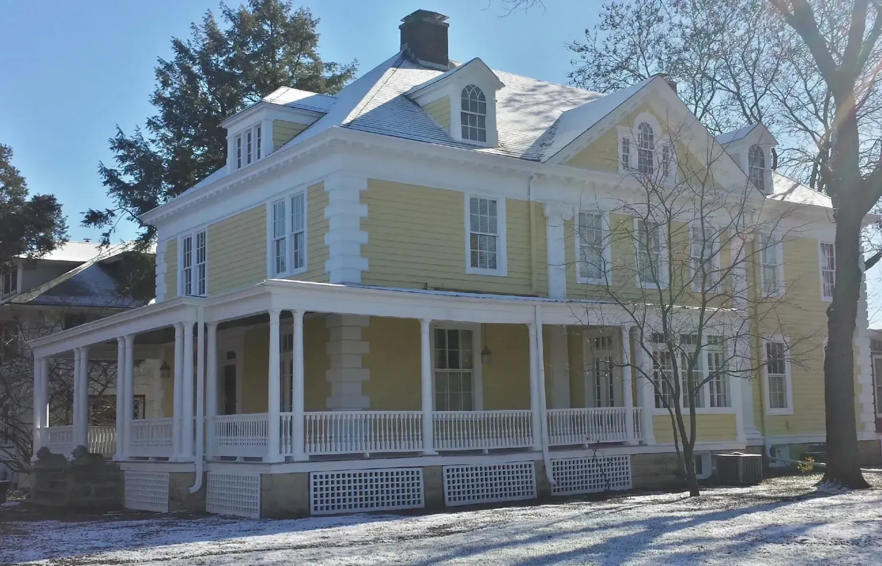 Large yellow Victorian-style house with a wraparound porch and white trim in a snowy yard