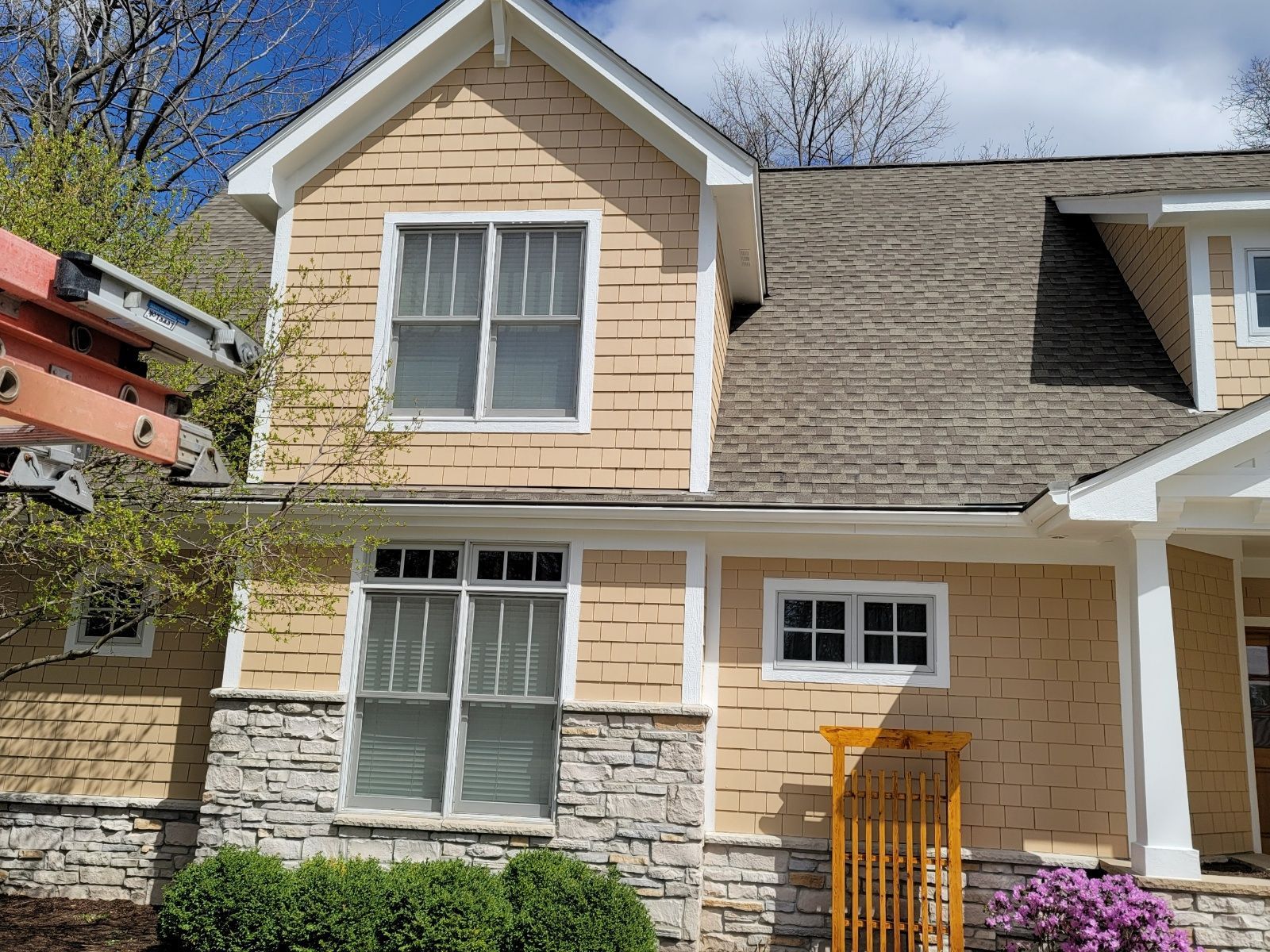 Two-story beige house with stone siding, white-trimmed windows, and a ladder at left.