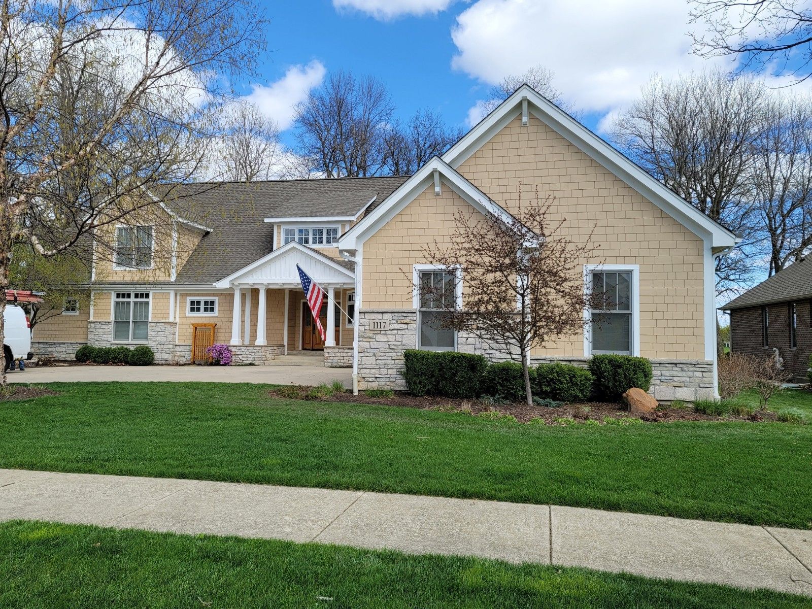 Suburban house with beige siding, stone accents, white columns, and a front lawn under a blue sky.