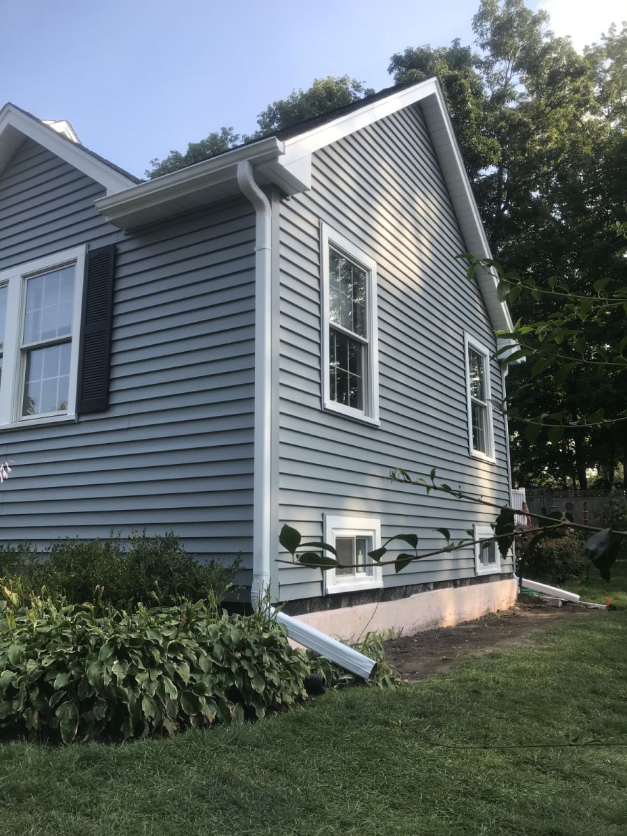 Gray vinyl-sided house exterior with windows, shrubs, and a sloped roof in a grassy yard