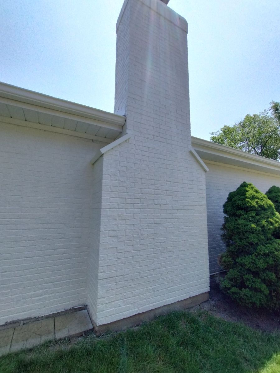 White brick exterior wall with a chimney and trimmed shrubs beside a grassy lawn