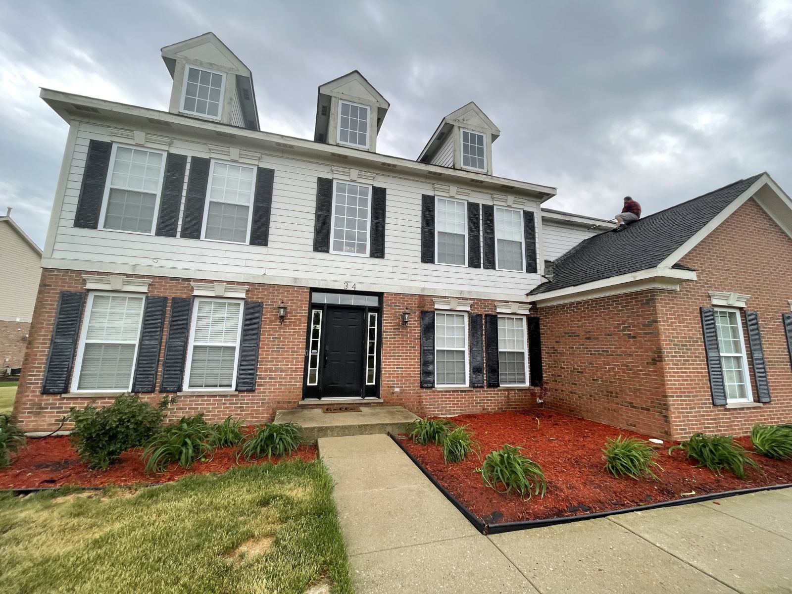 Three-story white and brick house with dormer windows, black shutters, and a red mulch front yard.