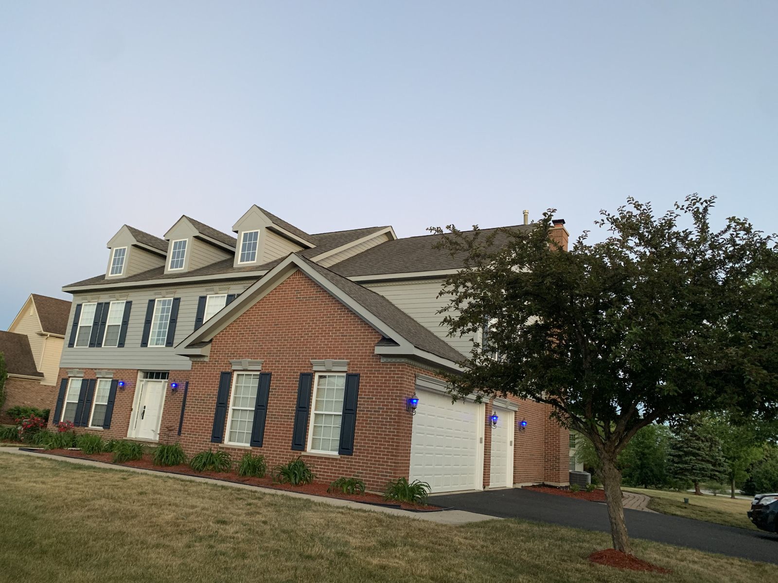 Two-story suburban house with brick and beige siding, blue shutters, a garage, and a front tree at dusk