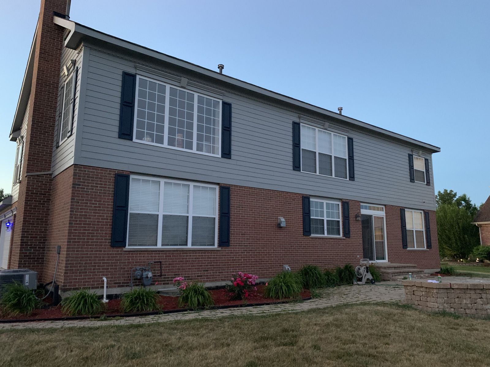 Two-story brick and gray house with white shutters and a small flower bed under a clear sky