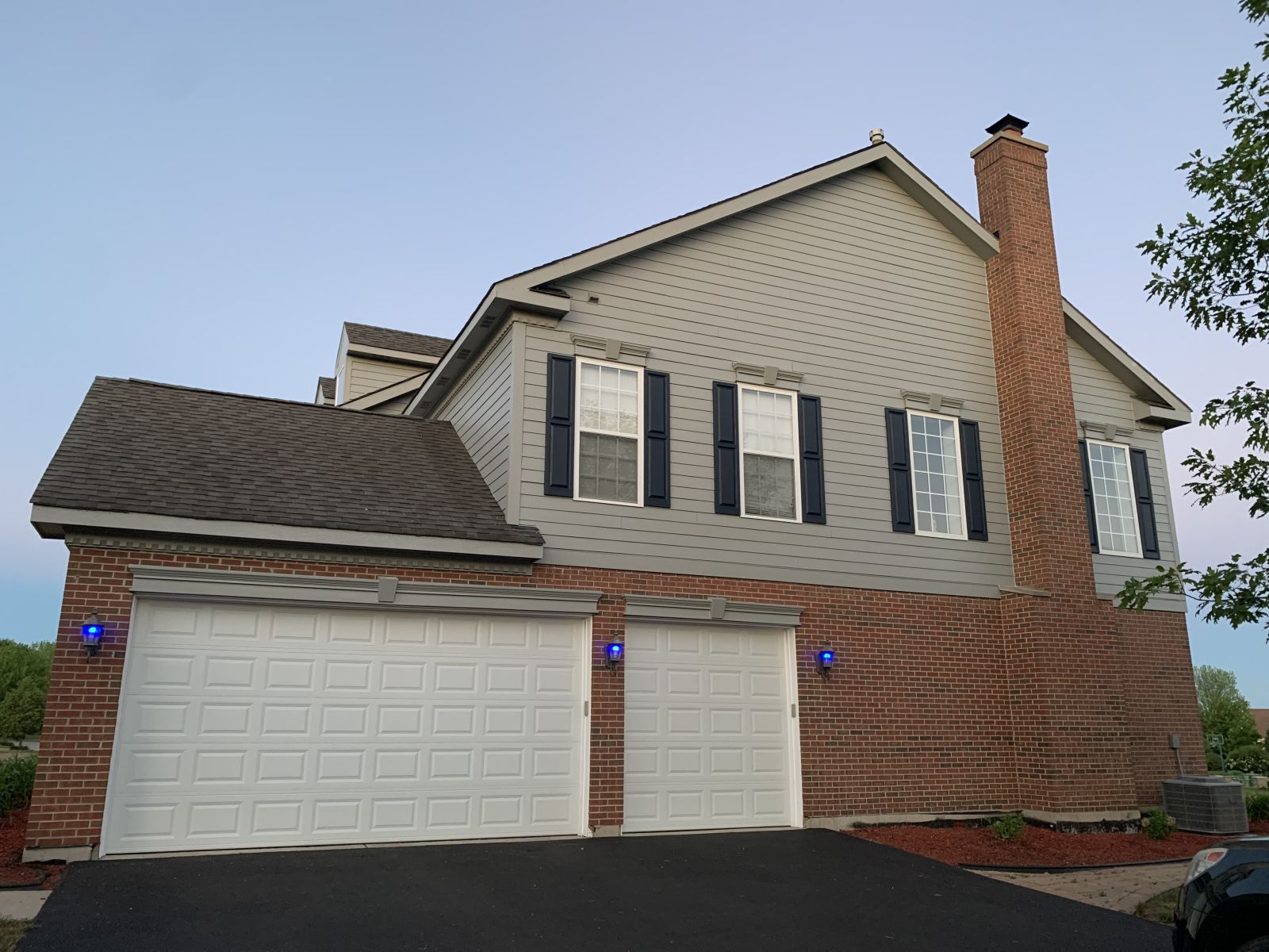 Suburban two-story brick house with white garage doors, black shutters, and a tall brick chimney