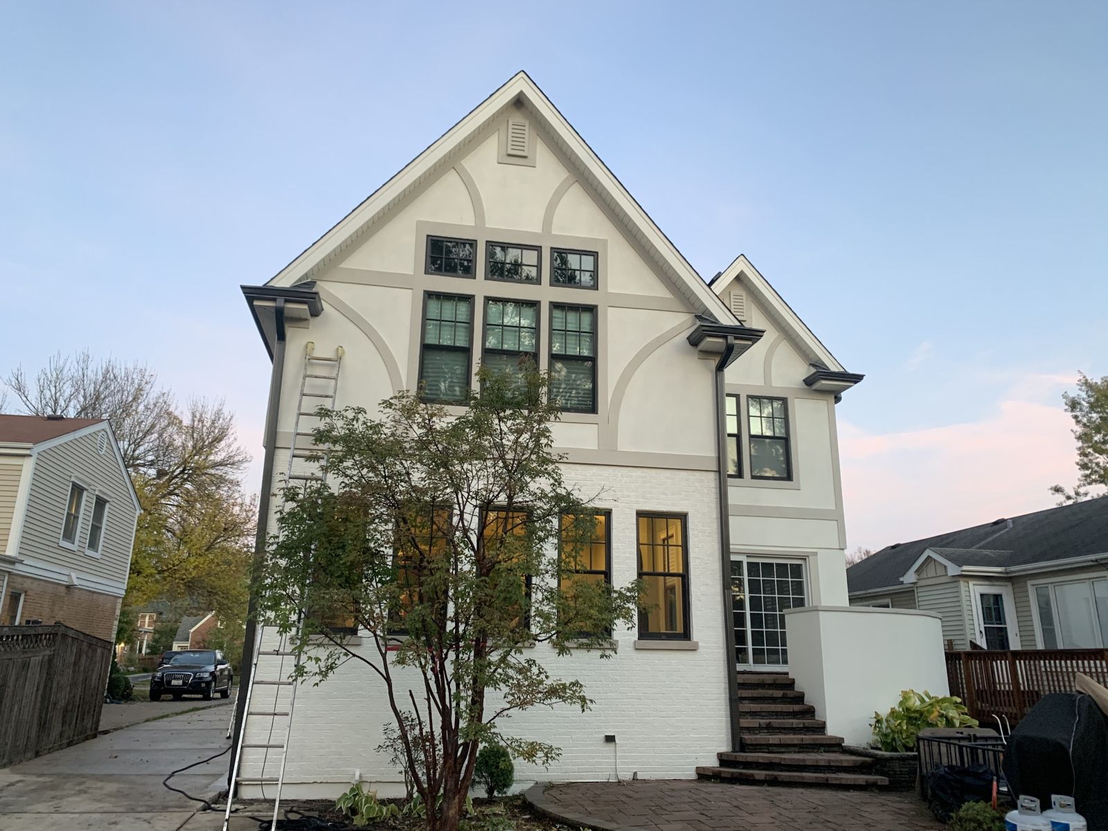 White two-story house with a steep roof, front tree, and parked cars on a residential street.