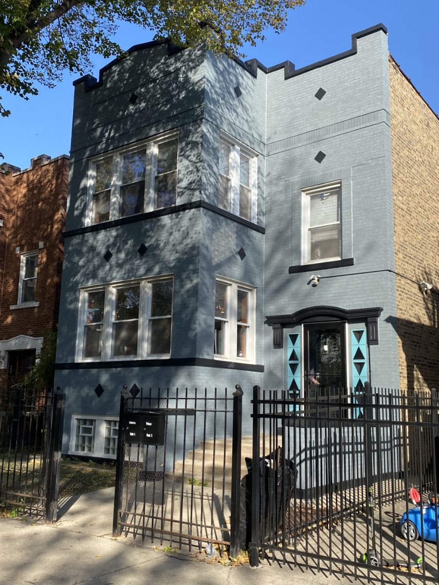 Gray three-story townhouse with black iron fence and gate, sunlit facade, and front steps.