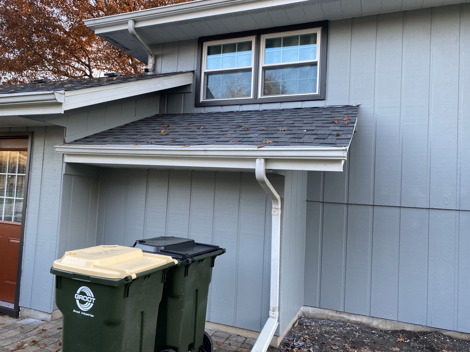 Two garbage bins beneath a small white awning attached to a gray house.
