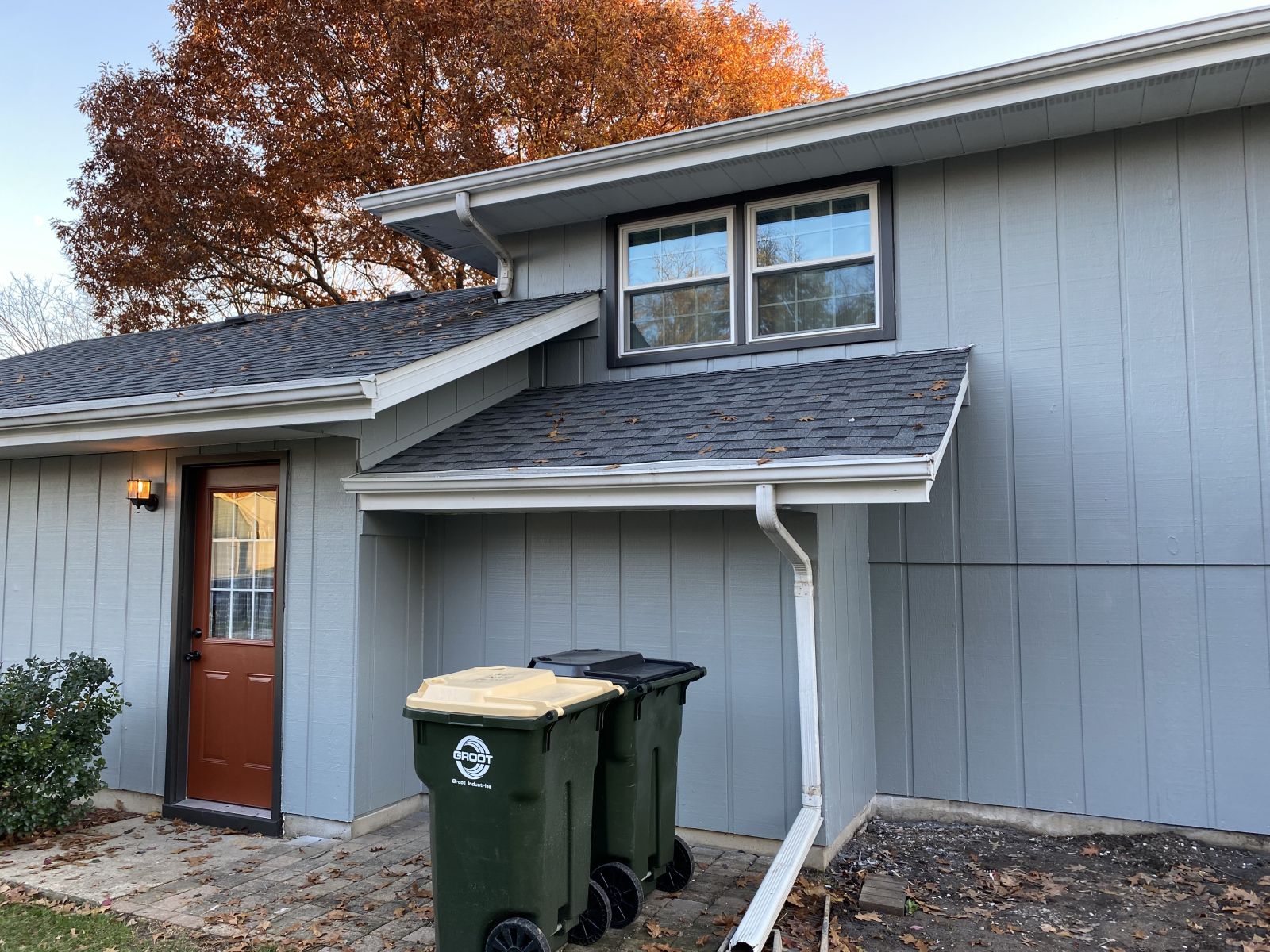 Two-story gray house with a red front door and trash bins near the entrance