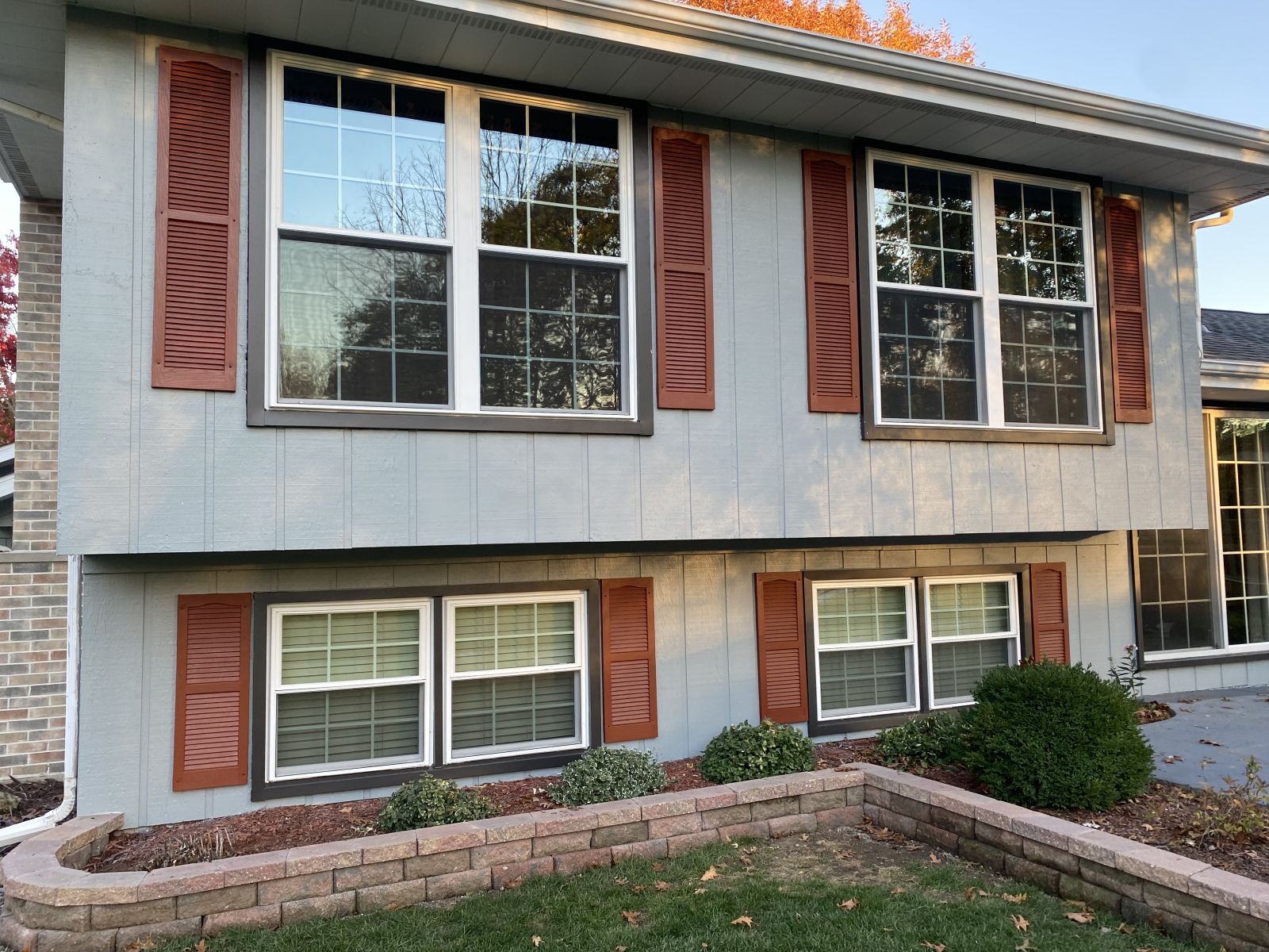 Two-story gray house with red shutters and windows, front shrub bed, and brick trim