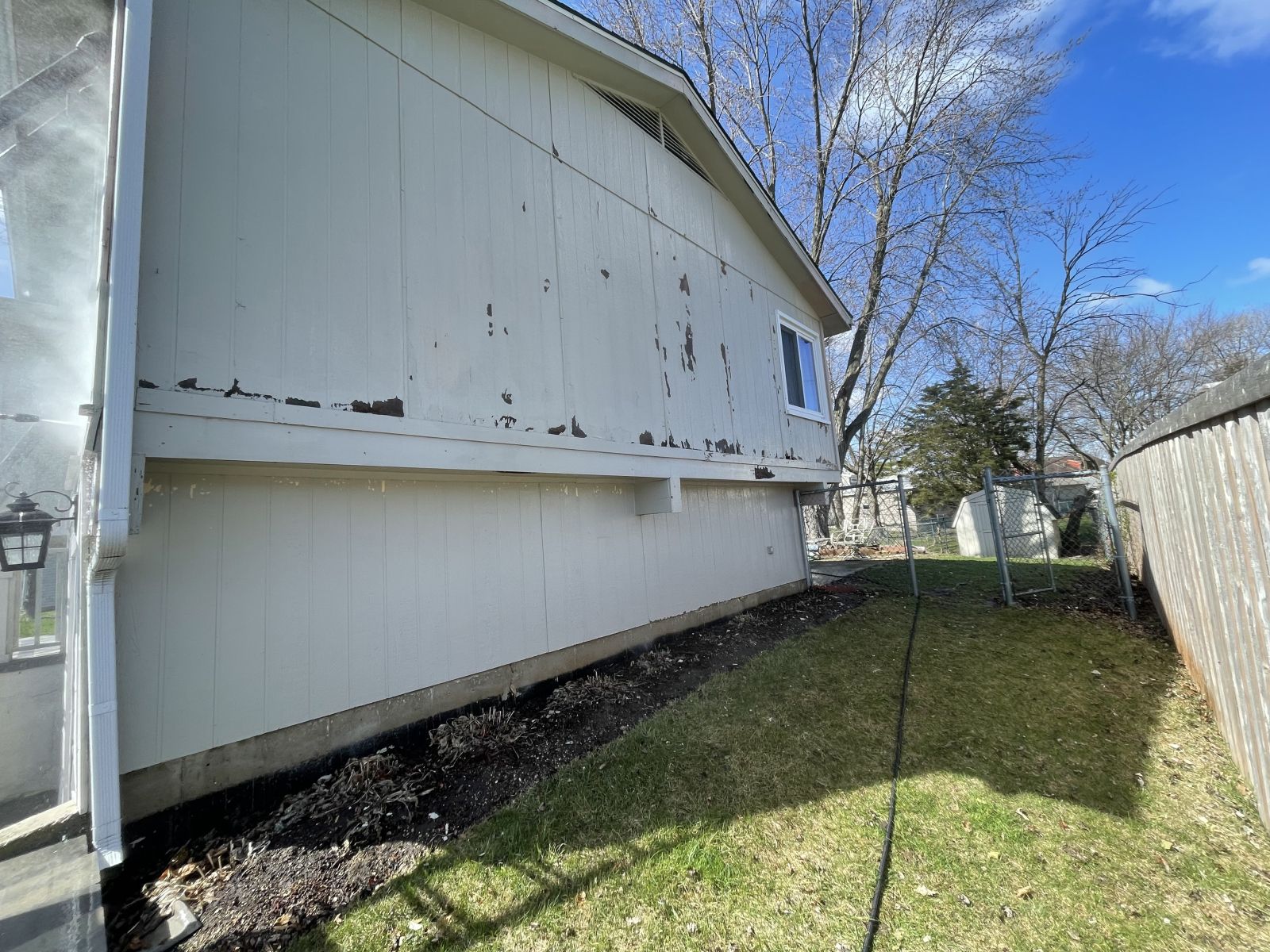 White mobile home siding with peeling paint and a small side window beside a grassy yard and fence.