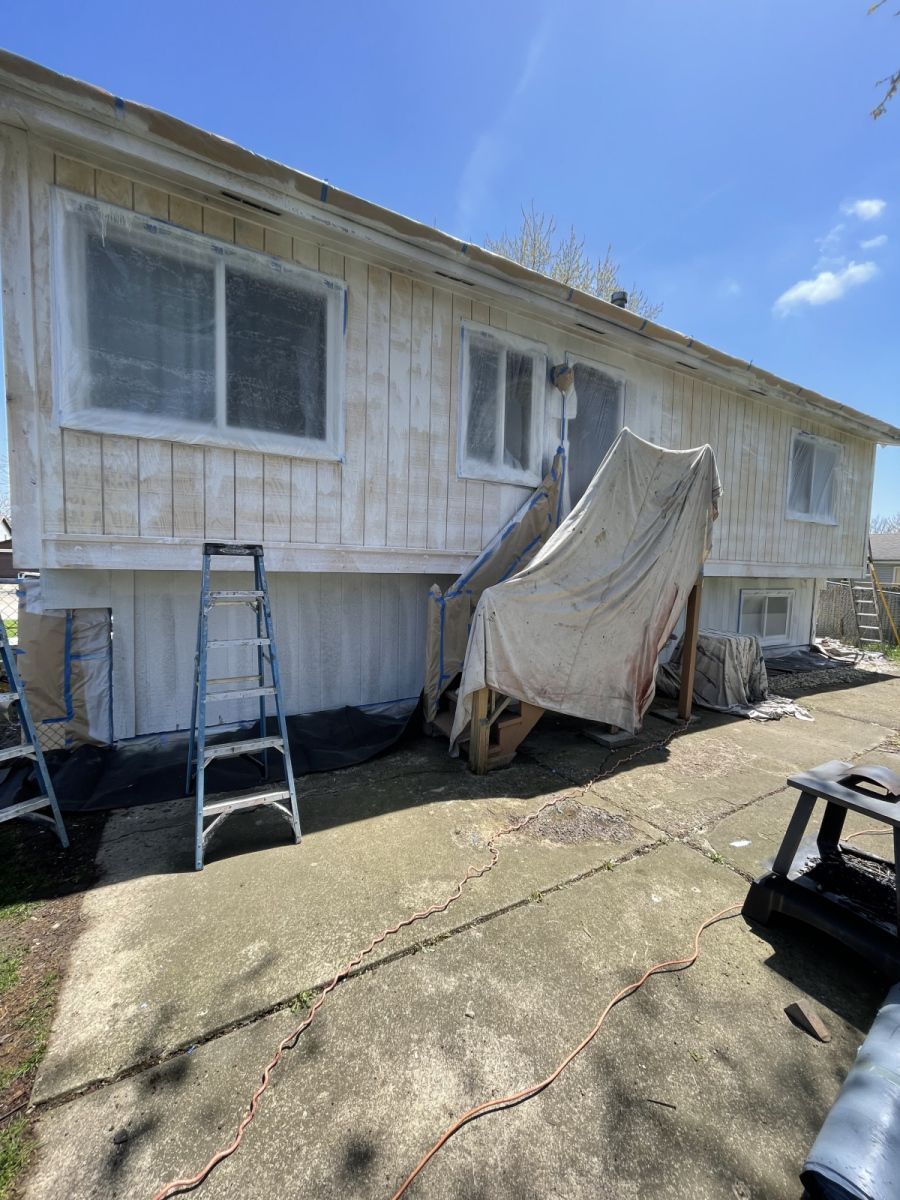 Damaged white mobile home with ladder and torn tarp draped over the porch in a sunny yard