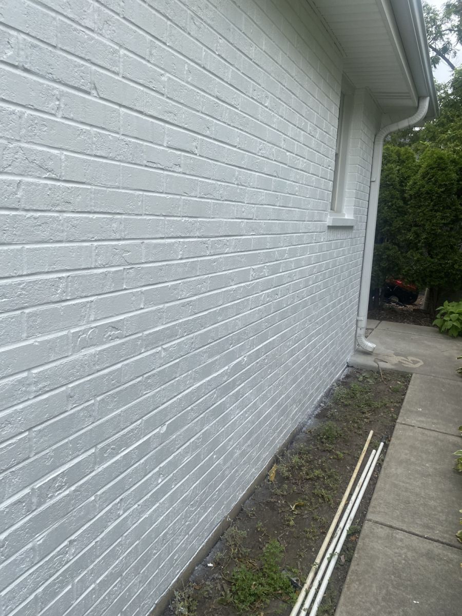 White brick house exterior beside a narrow concrete walkway and small garden bed with pipes.