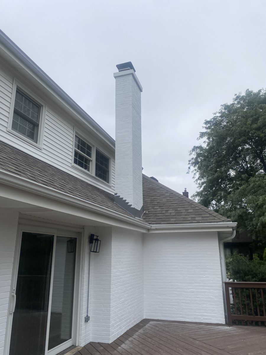 White house exterior with a brick chimney, sliding glass door, and shingled roof under an overcast sky