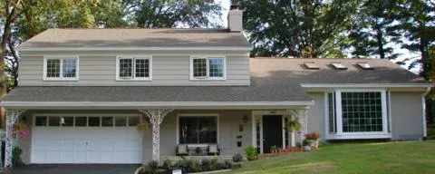 Suburban two-story house with white garage, porch, and large windows, surrounded by trees and lawn