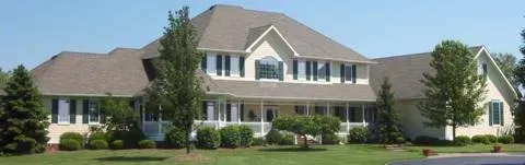 Large beige suburban house with multiple gables, white trim, and a manicured lawn on a sunny day