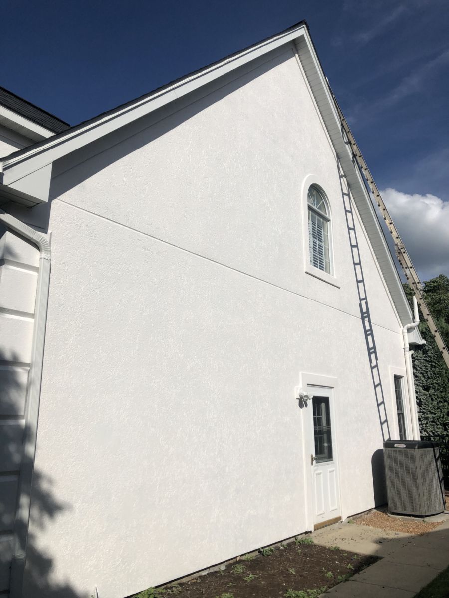 White house side wall with a steep roof, small windows, and an air-conditioning unit on the ground.