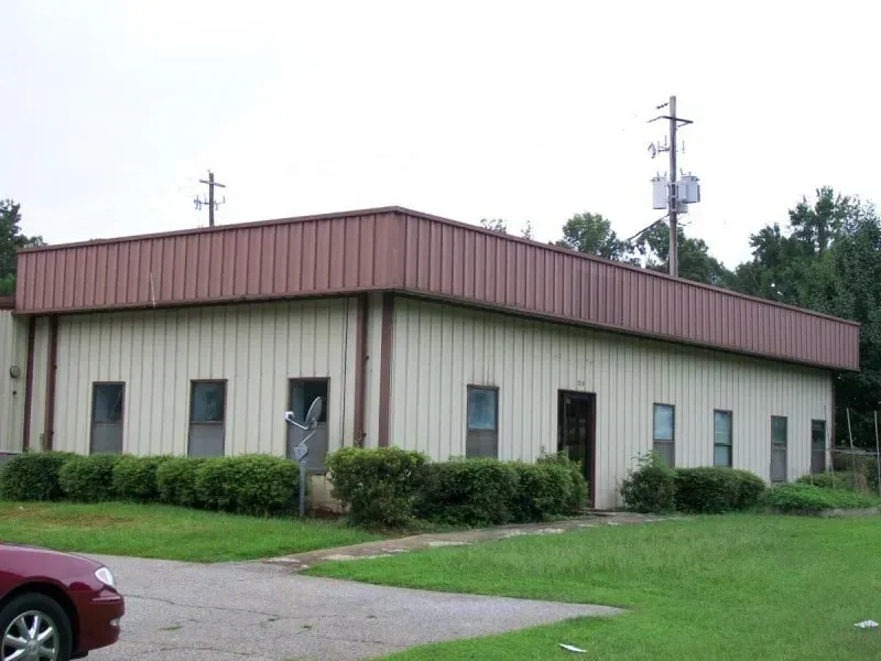 Single-story beige building with brown roof, small windows, and trimmed hedges beside a parking lot