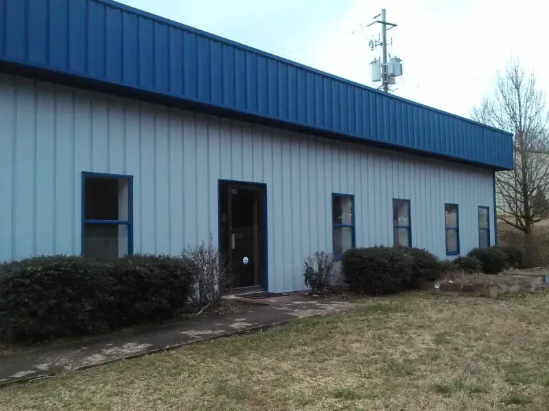 Small white industrial building with blue roof and windows, seen from outside with shrubs and grass.