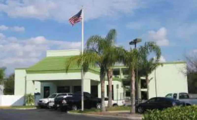 Light green building with an American flag, palm trees, and parked cars in front