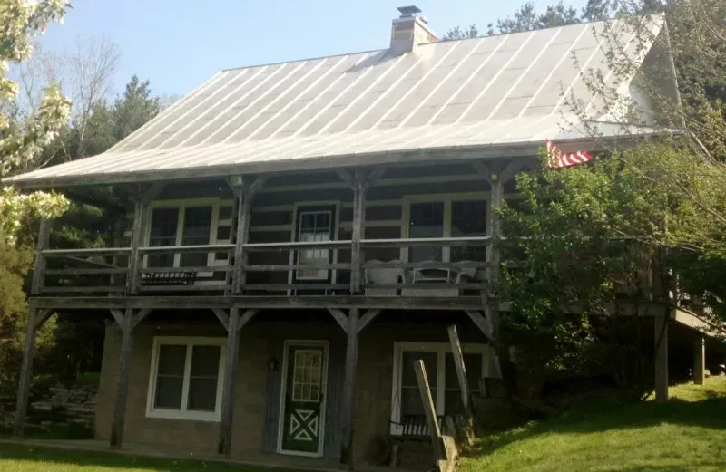 Two-story house with a white metal roof, front porch, and green trim surrounded by trees.