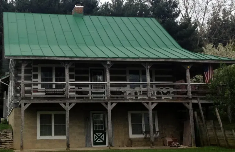 Two-story wooden house with green metal roof and porch, set among trees