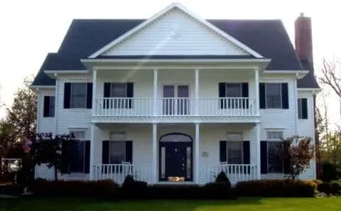 White two-story house with a front porch, balcony, and dark roof under a clear sky